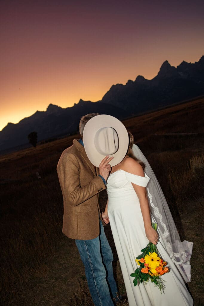 Bride and groom hide behind cowboy hat for a romantic kiss